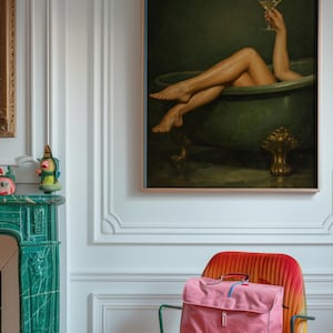 May include: A framed painting depicts a person relaxing in a green bathtub, holding a martini. A pink satchel bag sits on a red and orange chair. The room features white walls and a green fireplace.