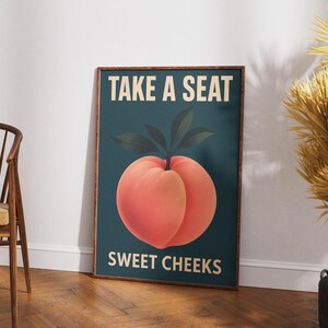 May include: A framed art print with a peach illustration and the text "TAKE A SEAT SWEET CHEEKS". The peach is orange-red with green leaves on a teal background. The brown frame complements the artwork. The print is on display.