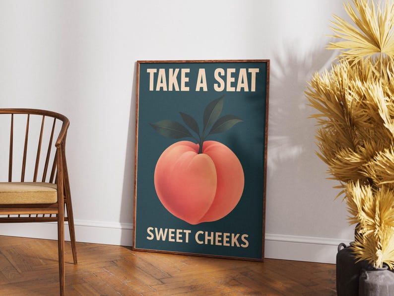 May include: A framed art print with a peach illustration and the text "TAKE A SEAT SWEET CHEEKS". The peach is orange-red with green leaves on a teal background. The brown frame complements the artwork. The print is on display.