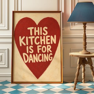 Könnte beinhalten: Gerahmter Kunstdruck mit einem großen roten Herz und dem cremefarbenen Schriftzug "THIS KITCHEN IS FOR DANCING". Das Kunstwerk befindet sich in einem goldenen Rahmen und wird vor einer weißen Wand präsentiert. Der Boden hat ein blau-weißes Schachbrettmuster.