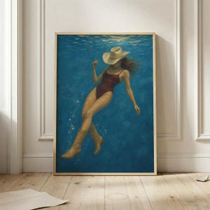 May include: A framed artwork depicts a person submerged in blue water, wearing a red swimsuit, cowboy boots, and a beige cowboy hat. The artwork has a wooden frame and is displayed on a wooden floor.