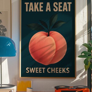 May include: A framed poster featuring a peach illustration and the text "TAKE A SEAT SWEET CHEEKS". The peach is a vibrant orange-red colour with green leaves against a dark teal background. The poster is in a gold frame and is displayed on a wall.