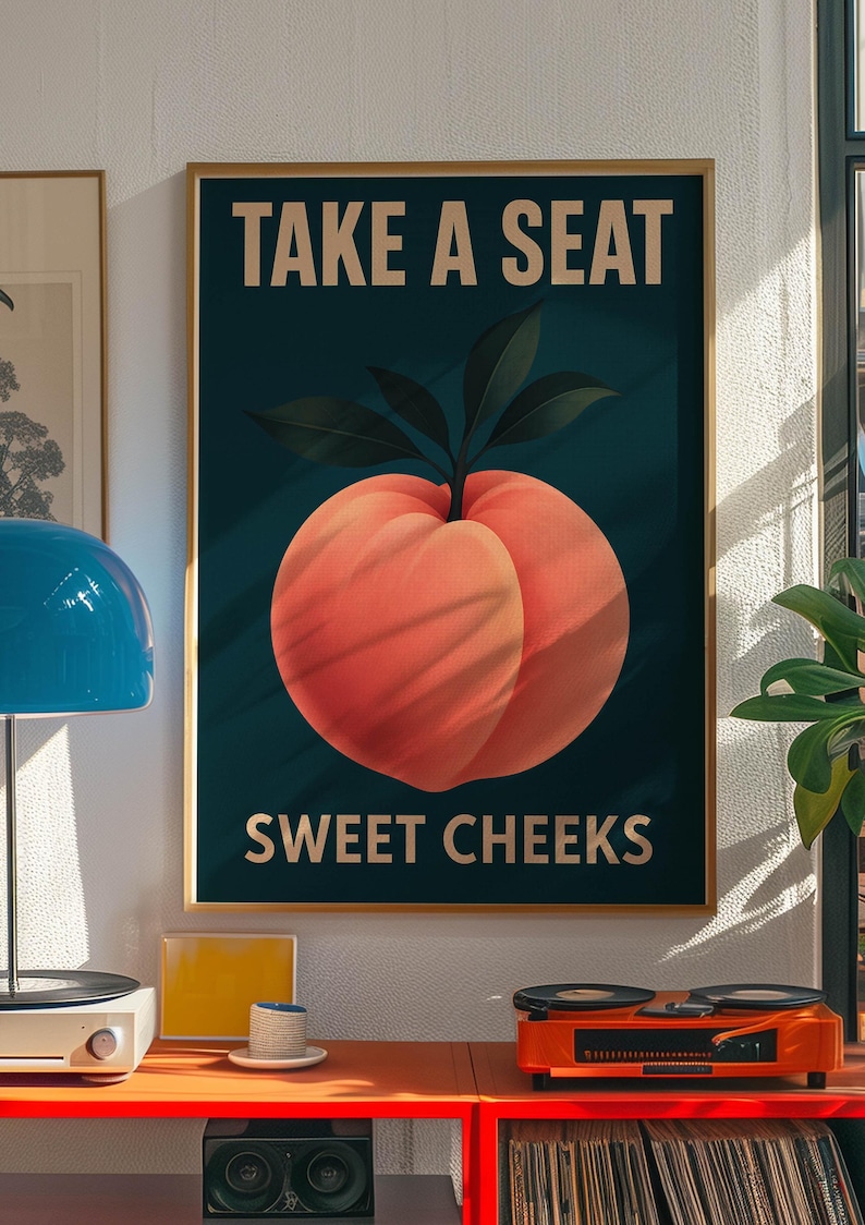 May include: A framed poster featuring a peach illustration and the text "TAKE A SEAT SWEET CHEEKS". The peach is a vibrant orange-red colour with green leaves against a dark teal background. The poster is in a gold frame and is displayed on a wall.
