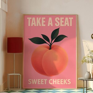 Puede incluir: Un p&oacute;ster rosa con el texto "TAKE A SEAT" y "SWEET CHEEKS" presenta una gran ilustraci&oacute;n de melocot&oacute;n. El melocot&oacute;n es de color naranja vibrante con hojas verdes. El p&oacute;ster est&aacute; en un marco dorado.