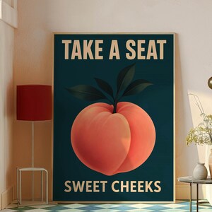 May include: A framed art print with a large peach illustration. The text "TAKE A SEAT SWEET CHEEKS" is in white on a dark teal background. The peach is a vibrant orange-red colour with green leaves. The artwork is a humorous piece.