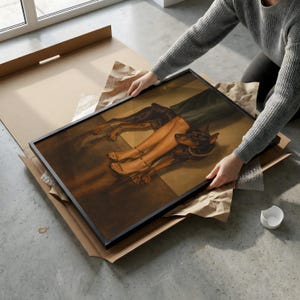 May include: A framed artwork featuring a Doberman Pinscher with a gold collar, lying near a pair of legs in sandals. The artwork is in a black frame, being removed from a cardboard box.