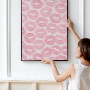May include: A framed artwork featuring a pattern of pink lipstick kiss prints on a light pink background. The black frame contrasts with the soft colors of the artwork. The artwork is being held up against a white wall.