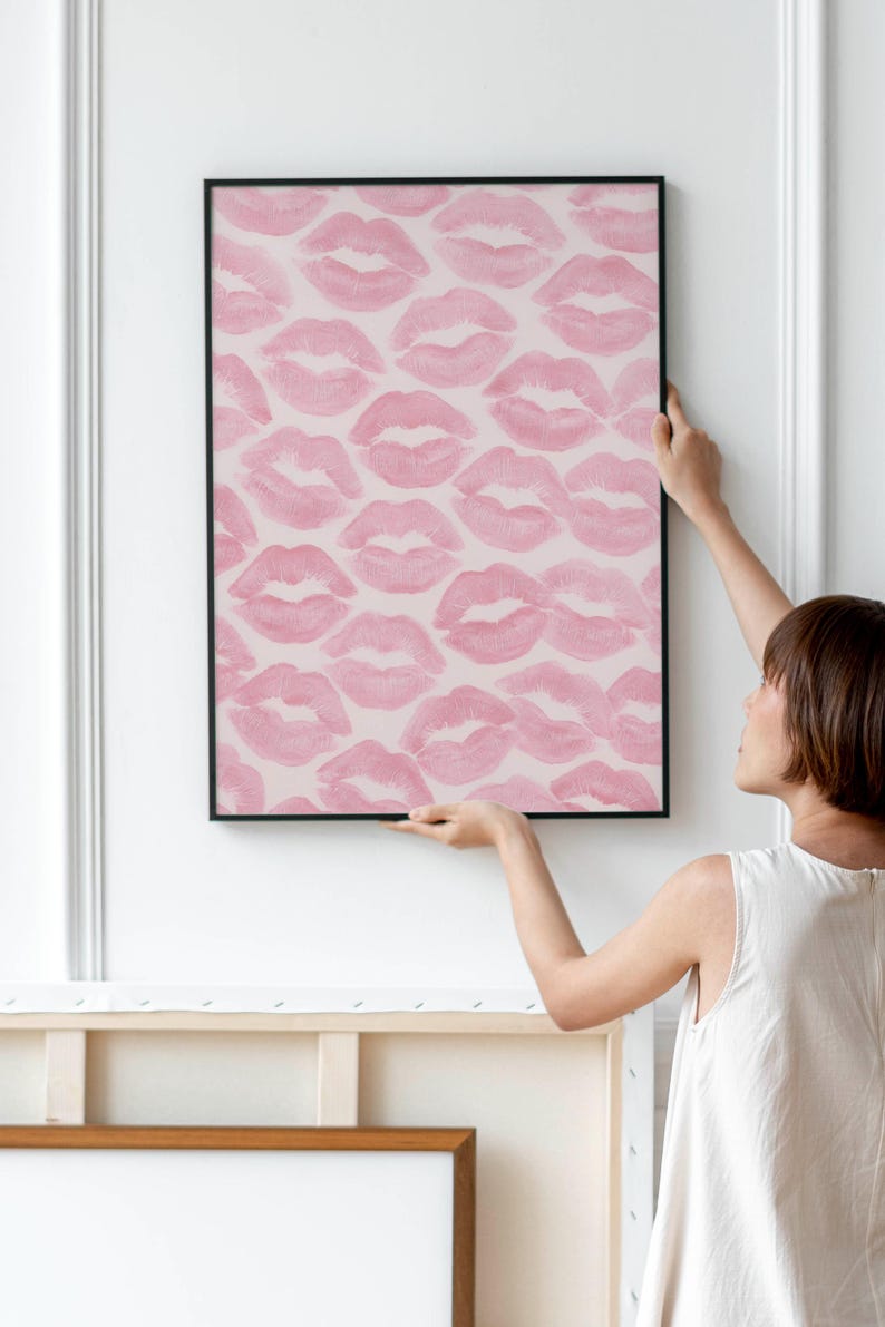 May include: A framed artwork featuring a pattern of pink lipstick kiss prints on a light pink background. The black frame contrasts with the soft colors of the artwork. The artwork is being held up against a white wall.