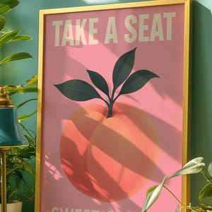 Puede incluir: Un cuadro enmarcado con fondo rosa e ilustraci&oacute;n de melocot&oacute;n. El texto "TAKE A SEAT" y "SWEET CHEEKS" est&aacute; impreso en blanco. El marco es dorado.