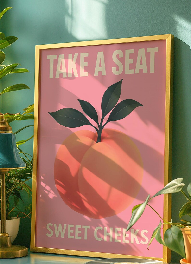 Puede incluir: Un cuadro enmarcado con fondo rosa e ilustraci&oacute;n de melocot&oacute;n. El texto "TAKE A SEAT" y "SWEET CHEEKS" est&aacute; impreso en blanco. El marco es dorado.