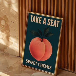 May include: A framed art print with a gold border. The artwork features a peach illustration on a dark teal background, with the text "TAKE A SEAT" and "SWEET CHEEKS" in white. The print is leaning against a wooden wall.