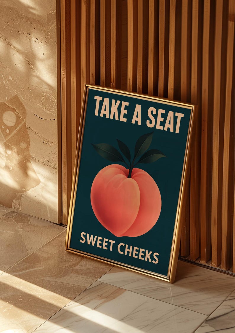 May include: A framed art print with a gold border. The artwork features a peach illustration on a dark teal background, with the text "TAKE A SEAT" and "SWEET CHEEKS" in white. The print is leaning against a wooden wall.