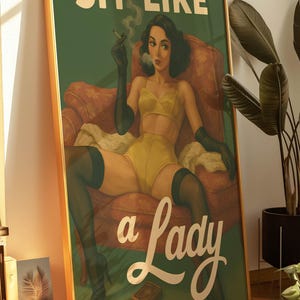 May include: Framed vintage-style poster with a woman in yellow lingerie, black gloves, and stockings, smoking a cigarette while seated in a red armchair. The text "SIT LIKE a Lady" is displayed in gold lettering on a green background.