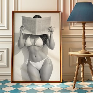 May include: A framed monochrome photograph of a person in a white bikini, holding a newspaper in front of their face. The photograph is in a gold frame and is leaning against a white wall. A pencil is held near the newspaper.
