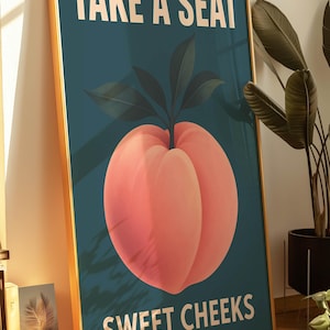 May include: Framed art print featuring a large peach illustration on a teal background. The peach is pink with green leaves. White text reads "TAKE A SEAT" and "SWEET CHEEKS". The frame has a gold finish.