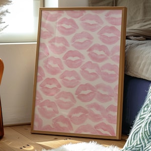 May include: A framed artwork featuring a pattern of pink lipstick kiss prints on a cream background. The wooden frame is rectangular and leans against a wall. The artwork is placed on a wooden floor with a white fluffy rug.