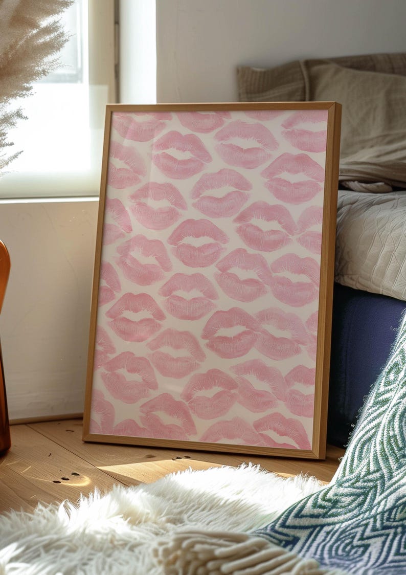 May include: A framed artwork featuring a pattern of pink lipstick kiss prints on a cream background. The wooden frame is rectangular and leans against a wall. The artwork is placed on a wooden floor with a white fluffy rug.
