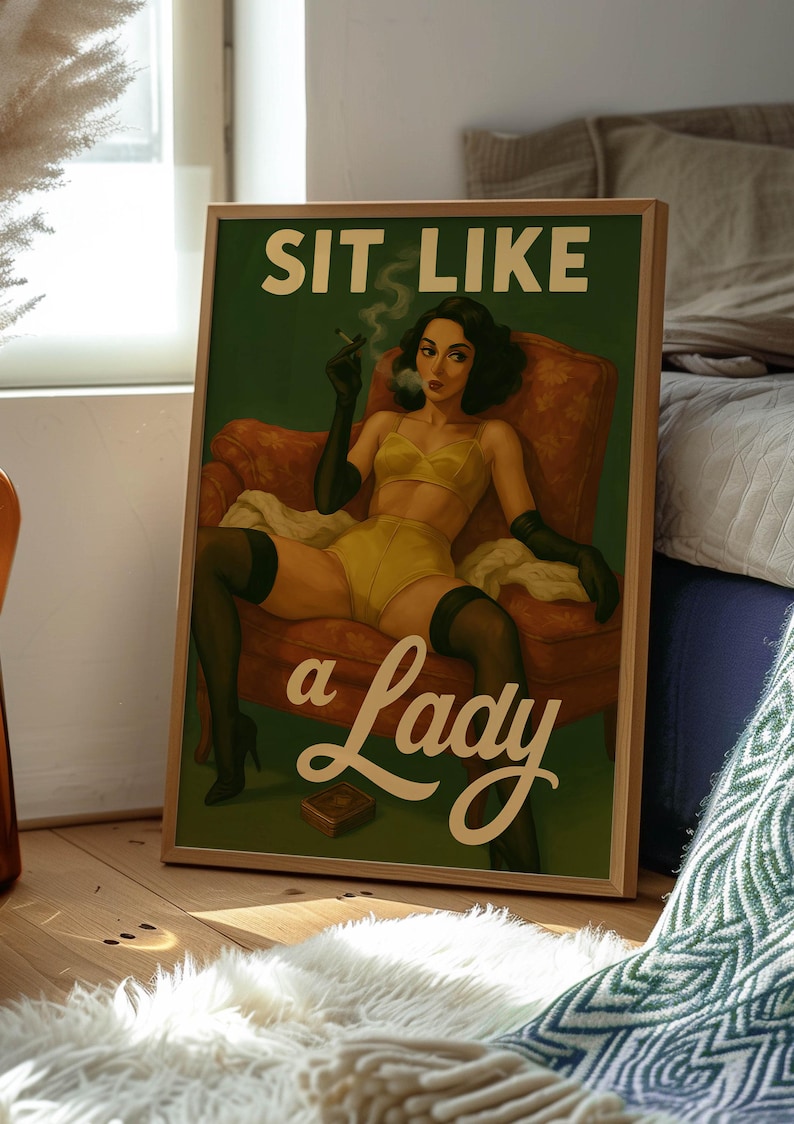 May include: Framed vintage-style poster with the text "SIT LIKE a Lady." The artwork depicts a woman in yellow lingerie, black gloves, and stockings, smoking a cigarette while seated in a red armchair. The poster is in a wooden frame.