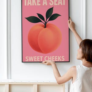 Puede incluir: Una impresi&oacute;n art&iacute;stica enmarcada con una ilustraci&oacute;n de melocot&oacute;n sobre un fondo rosa. El melocot&oacute;n tiene hojas verdes y el texto "TAKE A SEAT SWEET CHEEKS" en beige claro. La obra de arte se muestra en una pared blanca.