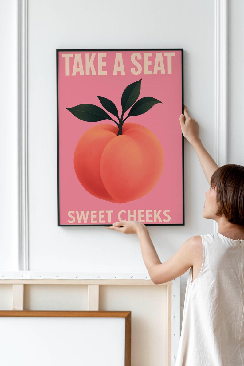 Puede incluir: Una impresi&oacute;n art&iacute;stica enmarcada con una ilustraci&oacute;n de melocot&oacute;n sobre un fondo rosa. El melocot&oacute;n tiene hojas verdes y el texto "TAKE A SEAT SWEET CHEEKS" en beige claro. La obra de arte se muestra en una pared blanca.