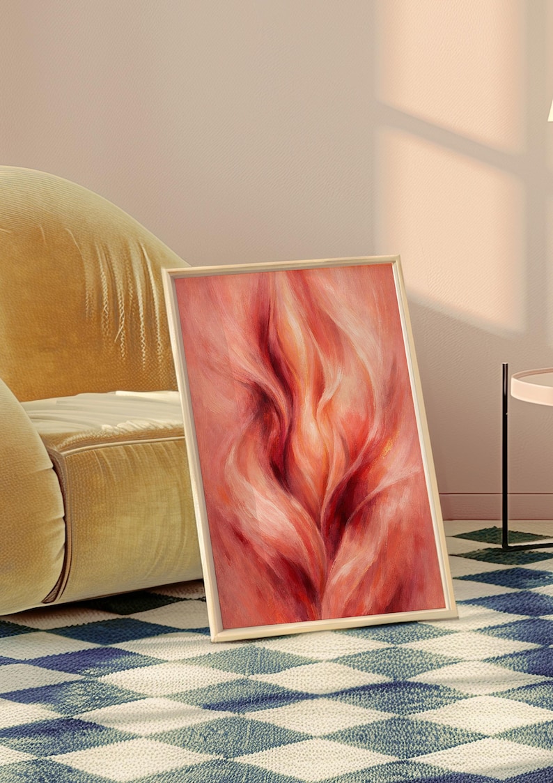 May include: A framed abstract art print with a flowing, flame-like design in red and peach hues. The artwork is displayed on a blue and white diamond patterned rug, leaning against a yellow velvet armchair.