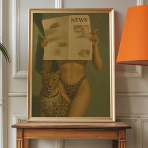 May include: Framed artwork featuring a woman holding a newspaper with the headline "NEWS", a leopard, and a cigarette. The woman wears leopard print underwear. The artwork is in a gold frame and sits on a wooden table.