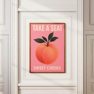 Puede incluir: Un cuadro enmarcado con una ilustraci&oacute;n de melocot&oacute;n sobre fondo rosa. El melocot&oacute;n es de color naranja con hojas verdes. El texto "TAKE A SEAT" y "SWEET CHEEKS" se muestra en blanco. El cuadro est&aacute; en un marco de madera.