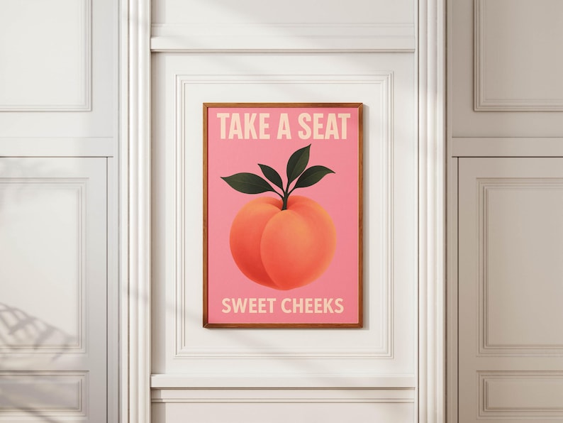 Puede incluir: Un cuadro enmarcado con una ilustraci&oacute;n de melocot&oacute;n sobre fondo rosa. El melocot&oacute;n es de color naranja con hojas verdes. El texto "TAKE A SEAT" y "SWEET CHEEKS" se muestra en blanco. El cuadro est&aacute; en un marco de madera.