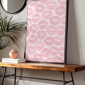 May include: A framed art print featuring a pattern of pink lipstick kiss marks on a white background. The artwork is displayed on a wooden bench with black metal legs, alongside a decorative vase and books. A round mirror hangs on the wall.