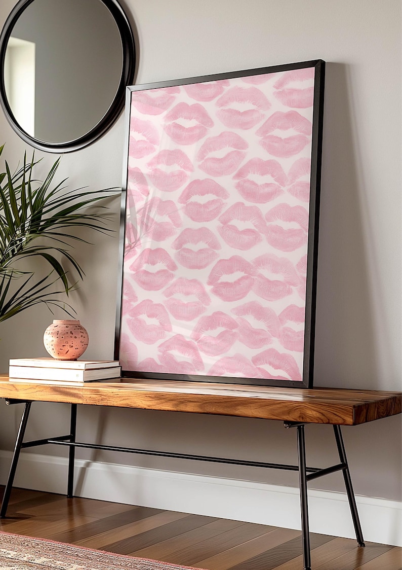 May include: A framed art print featuring a pattern of pink lipstick kiss marks on a white background. The artwork is displayed on a wooden bench with black metal legs, alongside a decorative vase and books. A round mirror hangs on the wall.