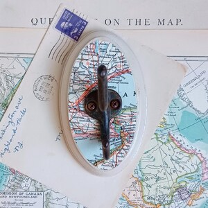 May include: A white ceramic hook with a map of Ontario, Canada printed on it. The hook is shaped like an oval and has a black metal hook attached to it. The map is in shades of blue, green, red, and yellow.