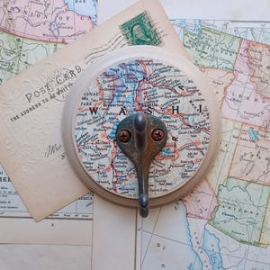 May include: A vintage-style wooden hook with a metal hook, featuring a map of Washington state with the text "WASH" and "Seattle" on the front. The hook is surrounded by vintage maps.