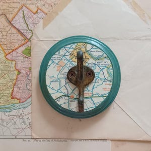 May include: A teal blue round wall hook with a map of Philadelphia, Pennsylvania printed on the front. The hook is made of metal and has a rustic finish.