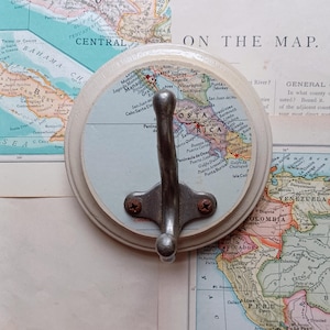 May include: A white ceramic hook with a silver metal hook. The hook is decorated with a vintage map of Costa Rica. The map is blue and white with brown text. The hook is on a white background.