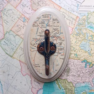 May include: A white oval-shaped wall hook with a blue metal hook. The hook is decorated with a map of Arizona with the city of Phoenix highlighted. The hook is mounted on a map of North America.