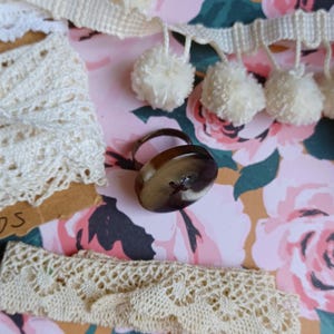 May include: A brown and white button ring with a metal band. The ring is on a floral background with white pom-poms.