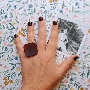 May include: A hand wearing a large, square, brown ring with a gold accent. The hand is painted with dark brown nail polish.