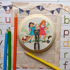 May include: A vintage, cream-colored, round, metal, wall-mounted hook with a metal ring. The hook features a colorful illustration of two girls and a dog on roller skates. The text "THE UNITED" is visible in the background.