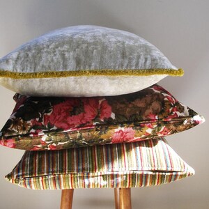 May include: A stack of three decorative throw pillows. The top pillow is a light gray velvet with a gold trim. The middle pillow is a brown and pink floral print. The bottom pillow is a striped fabric with green, brown, and white stripes.