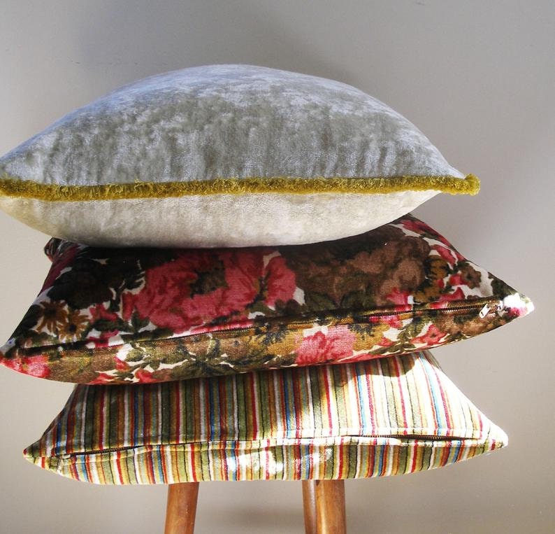 May include: A stack of three decorative throw pillows. The top pillow is a light gray velvet with a gold trim. The middle pillow is a brown and pink floral print. The bottom pillow is a striped fabric with green, brown, and white stripes.