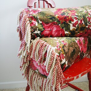 May include: A red wooden chair with a floral patterned pillow with fringe. The pillow is made of a soft, plush fabric and has a vintage look.