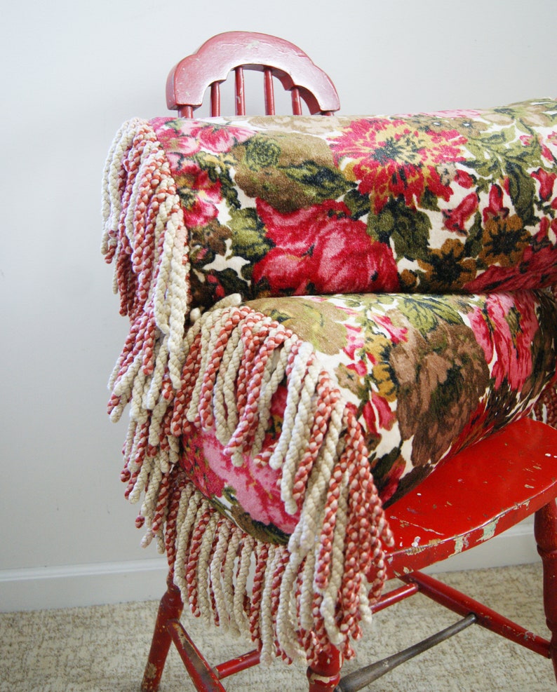 May include: A red wooden chair with a floral patterned pillow with fringe. The pillow is made of a soft, plush fabric and has a vintage look.