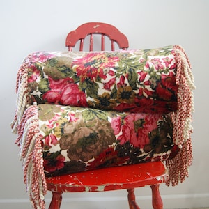 May include: Two floral patterned throw pillows with fringe trim, stacked on a red wooden chair.