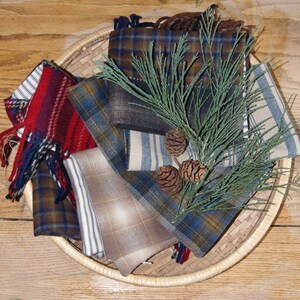 Puede incluir: Una colecci&oacute;n de muestras de tela a cuadros y a rayas en tonos de azul, marr&oacute;n, rojo y blanco, dispuestas en una cesta de mimbre con una rama de con&iacute;fera y pi&ntilde;as.