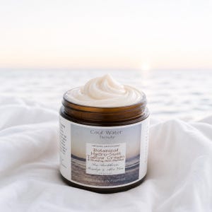 Może przedstawiać: Słoik kremu Botanical Hydro-Suet Tallow firmy Cool Water Beauty. Krem jest biały i zakręcony na wierzchu słoika. Słoik ma brązową pokrywkę i etykietę z tekstem. Tło to biała tkanina i ocean.