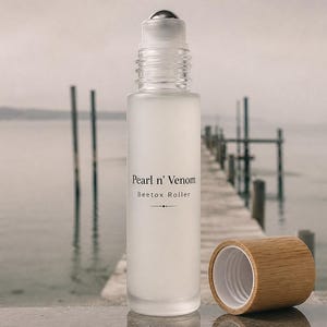 Op de afbeelding: Een matglazen rollerflesje met een zilveren rollerbal en een houten dop. Op de fles staat "Pearl n' Venom Beetox Roller". De dop is eraf, waardoor de opening van de fles zichtbaar is. De achtergrond toont een pier en water.