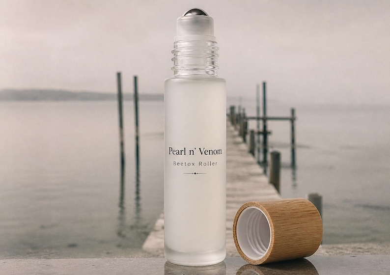 Op de afbeelding: Een matglazen rollerflesje met een zilveren rollerbal en een houten dop. Op de fles staat "Pearl n' Venom Beetox Roller". De dop is eraf, waardoor de opening van de fles zichtbaar is. De achtergrond toont een pier en water.