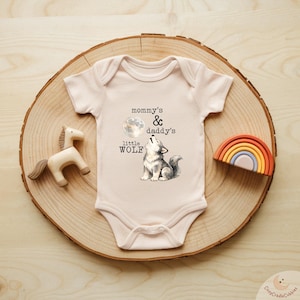 May include: A light pink baby bodysuit with the text "mommy's & daddy's little WOLF" and a graphic of a howling wolf under a full moon. A wooden horse and a rainbow-shaped toy are also present.
