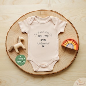 May include: A light pink baby onesie with the text "Aunt Ashley & Uncle Chris, WILL YOU BE MY Godparents?" is displayed on a wooden slice. A wooden horse and rainbow are also on the wooden slice.