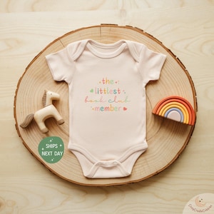 May include: A pink baby onesie with the text "the littlest book club member" in colorful letters. The onesie is on a wooden surface with a wooden toy horse and a rainbow stacking toy.